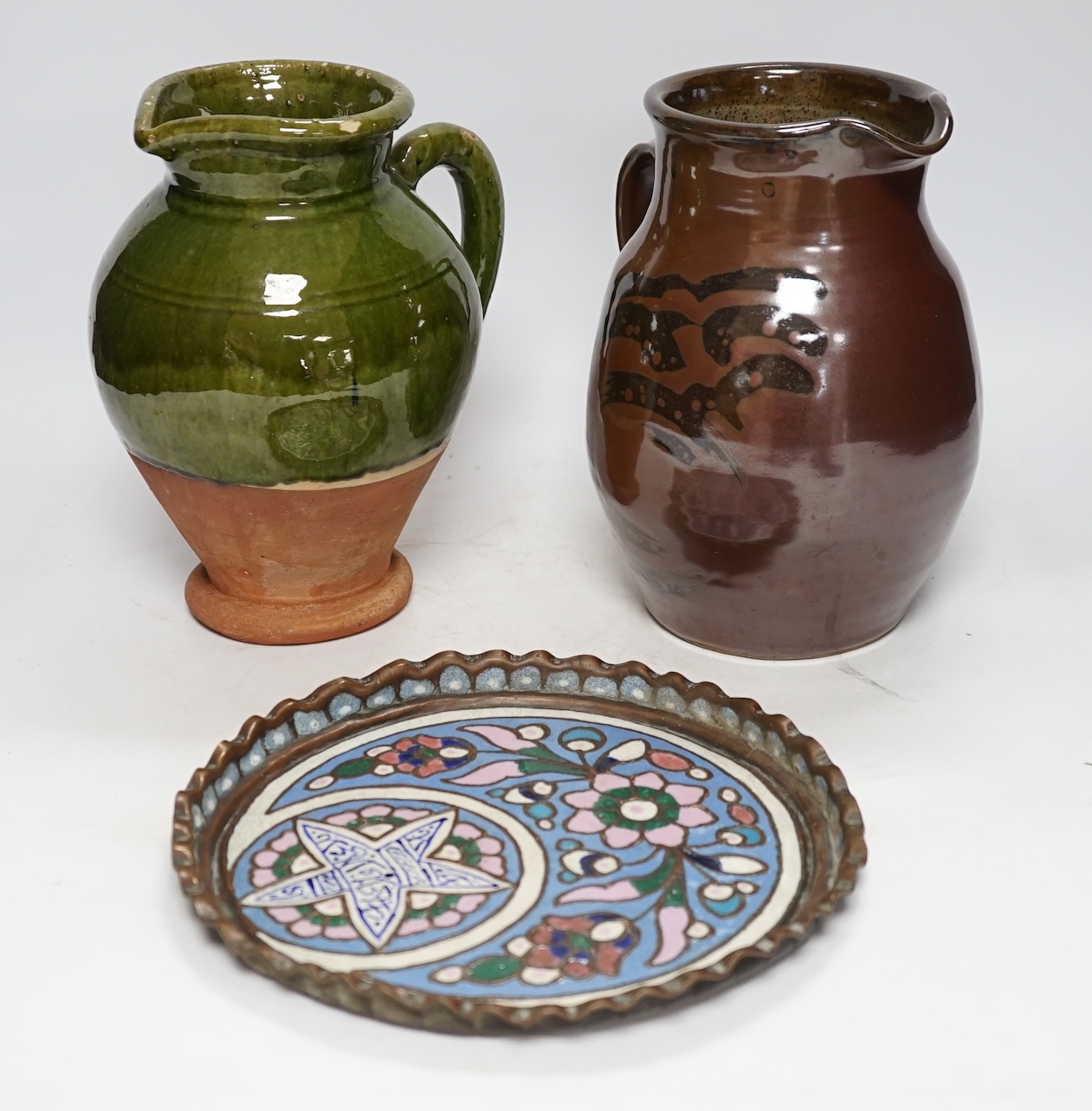 From the Studio of Fred Cuming. Two studio green and brown glazed jugs together with an enamelled stylised floral dish, largest height 22cm. Condition - good